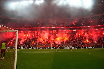 Vista de un estadio de fútbol lleno de aficionados con bengalas rojas y humo. El campo verde, una portería y varios jugadores son visibles en primer plano