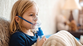 Niña rubia sentada con una mascarilla de nebulizador en el rostro mientras sostiene la pieza plástica con la mano.
