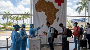 Pessoal médico usando equipamento de proteção individual mede a temperatura dos passageiros em um aeroporto. Um mapa da África e as palavras “As Américas” podem ser vistos ao fundo.
