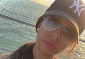 Primer plano de una persona con gorra negra de los Yankees y gafas de sol estilo aviador, con el mar de fondo bajo la luz del atardecer