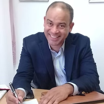 Hombre de mediana edad sonríe sentado en un escritorio con chaqueta azul marino y camisa clara a rayas, escribe con un bolígrafo en una oficina
