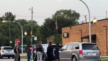 Oficiales de policía, civiles, vehículos SUV y miniván, edificio de ladrillo, bandera de Estados Unidos, cartel de stop y farolas en una calle