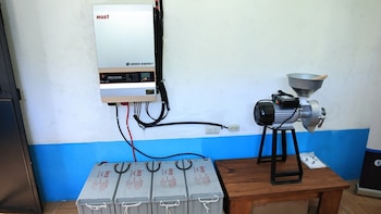 Inversor solar MUST GREEN ENERGY na parede, conectado a quatro baterias GOLD no chão e ao moinho elétrico na mesa de madeira.