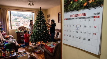 Dos mujeres y un niño decoran un árbol de Navidad junto a cajas de adornos. Un calendario de diciembre de dos mil veintiséis con una nota.