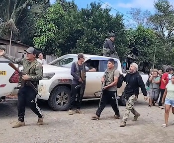Hombres armados, algunos con uniforme camuflado, junto a una camioneta blanca en un camino de tierra, con árboles y personas observando al fondo