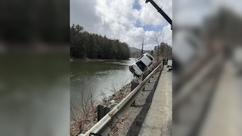 Una grúa elevando un coche blanco volcado de un río con orillas rocosas y árboles, junto a una carretera con barandilla. El cielo está parcialmente nublado