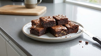 Primer plano de un plato blanco con brownies cuadrados de chocolate y nueces en una encimera gris. Un cuchillo dentado y migas están al lado del plato.
