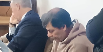 Un hombre con sudadera marrón y pelo oscuro, en primer plano de perfil, mira hacia abajo, mientras otra persona con traje oscuro se ve parcialmente detrás