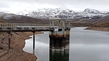 Torre de captación con pasarela en un embalse de agua, con una orilla de tierra y montañas nevadas al fondo.