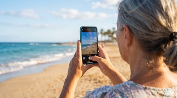 Una mujer vista de espaldas sostiene un teléfono móvil que enfoca una playa con mar, arena y palmeras.