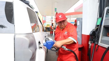 Un hombre con uniforme rojo de Petroperú sostiene una manguera azul de gasolina, llenando el tanque de un automóvil blanco en una estación de servicio