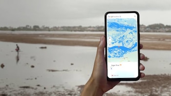 Una mano sostiene un teléfono inteligente que muestra la aplicación Google Flood Hub con un mapa del río Níger y una alerta roja. Al fondo, un paisaje fluvial y una persona