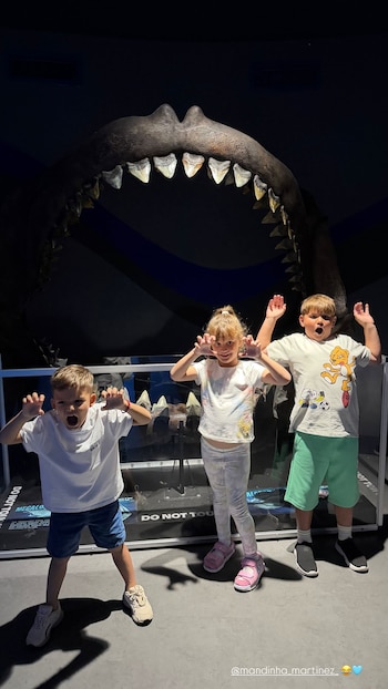 Tres niños posan frente a una exhibición de mandíbula de tiburón con dientes grandes y afilados y letreros que indican "DO NOT TOUCH"