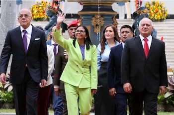 Varias personas con traje formal caminan al aire libre mientras una mujer con traje verde saluda con la mano levantada frente a una fuente
