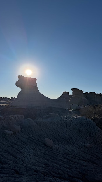 Un sol brillante se asoma detrás de una gran formación rocosa en un paisaje desértico. Hay otras rocas erosionadas y suelo agrietado