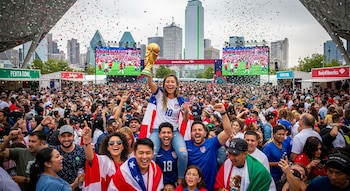 Una mujer sonriente con una camiseta de USA y un trofeo dorado, en hombros de un hombre, rodeada de una multitud jubilosa, banderas y confeti en Dallas.