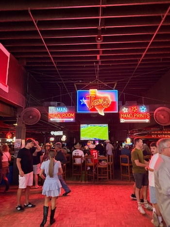 Interior de un local con techo de vigas, luz roja, personas de pie y sentadas, pantalla de televisión y carteles de neón con el texto "Texas"