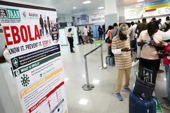 Un panel informativo sobre el ébola se ubica en primer plano ante una fila de viajeros con equipaje en la terminal de un aeropuerto
