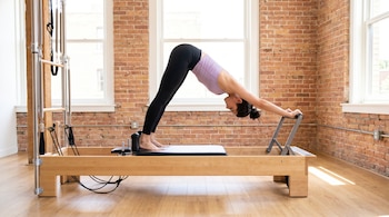 Mujer en ropa deportiva realiza estiramiento en reformador de Pilates de madera. Manos en la barra, pies en la plataforma. Fondo de ladrillo visto y ventanas.