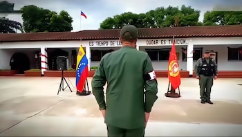 Edificio blanco con tejado en el Comando de Zona GNB Amazonas. Dos oficiales uniformados, banderas de Venezuela y otra roja, y un altavoz en el patio