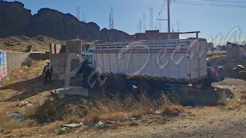 Un policía uniformado con rifle vigila un camión blanco y azul varado en un terreno sin pavimentar, con edificaciones y montañas al fondo bajo cielo claro