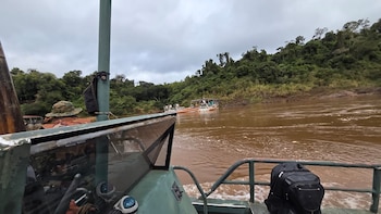 Vista desde un bote en un río. Aguas marrones y orillas con vegetación. Cielo nublado. Otro bote a lo lejos. Persona con sombrero militar y mochila negra en primer plano