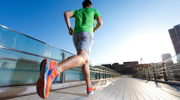 Un hombre con camiseta verde, pantalón corto gris y zapatillas de deporte naranja y azul corre en un puente. Hay barandas de vidrio y edificios.