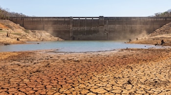 Suelo de tierra seca y agrietada en primer plano ante un pozo de agua y una estructura de concreto de una represa al fondo.