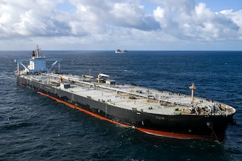 Vista aérea de un gran buque tanque petrolero negro con franja roja en el agua azul oscuro, bajo un cielo nublado, con otro barco pequeño al fondo