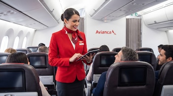 Tripulante de cabina de Avianca, vestida de rojo, interactúa con un pasajero en la cabina de un avión. Sostiene una tableta, con otros pasajeros sentados detrás.