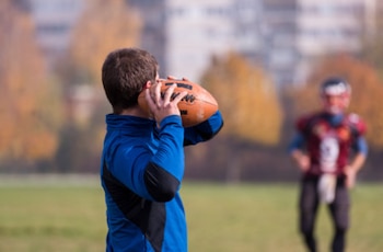 Joven de espaldas con camiseta azul, sujetando un balón de fútbol americano. Otro jugador con equipación de fútbol americano en un campo verde