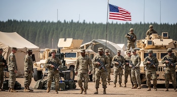 Grupo de soldados de EEUU con uniformes de camuflaje y fusiles, algunos de pie y otros sobre vehículos militares, en un campamento bajo la bandera estadounidense.
