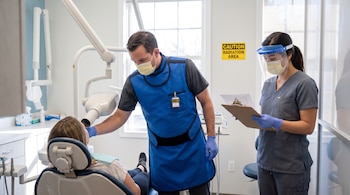 Dentista con mandil de plomo y mascarilla, asistente con protector facial y mascarilla. Paciente en sillón dental. Equipo de Rayos X y señal de radiación