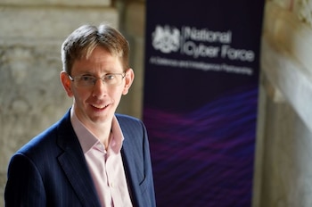 Un hombre con gafas y traje azul sonríe frente a un cartel con el texto National Cyber Force