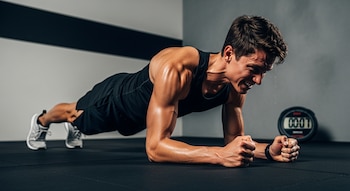 Hombre atlético realiza una plancha intensa en el suelo de un gimnasio, con expresión de esfuerzo y sudor. Un cronómetro digital desenfocado se ve a la derecha.