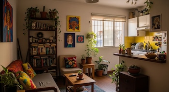 Vista interior de un departamento en CDMX: sofá, estantería con libros y plantas, arte en paredes, y cocina integrada con azulejos amarillos.