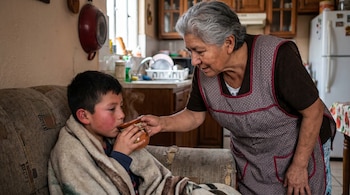 Una anciana con delantal da una taza humeante a un niño envuelto en una manta sobre un sofá, con una cocina de fondo.