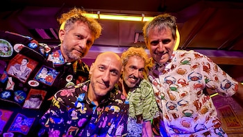 Cuatro hombres caucásicos posan sonrientes para la cámara. Visten camisas coloridas con estampados variados bajo luces de neón en un interior