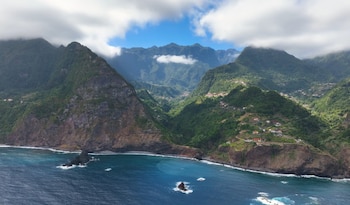 Vista aérea de una escarpada costa montañosa cubierta de vegetación, con casas dispersas en las laderas y un océano azul profundo con rocas en el agua