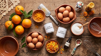 Ingredientes como naranjas, huevos, latas de leche, azúcar, ralladura, vainilla y pétalos sobre una mesa de madera con cuencos de barro.
