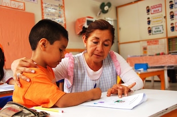 Una mujer adulta y un niño sentados frente a un escritorio. El niño escribe en una hoja, la mujer le asiste. Sobre el escritorio hay un estuche y lápices. Al fondo, un aula de clases