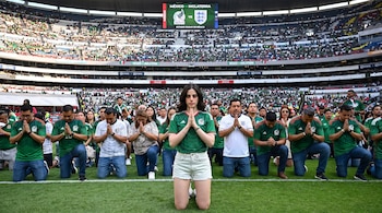 Grupo de personas arrodilladas en un campo de fútbol, con camisetas de México y una de Inglaterra, manos unidas, y un marcador electrónico en un estadio lleno.