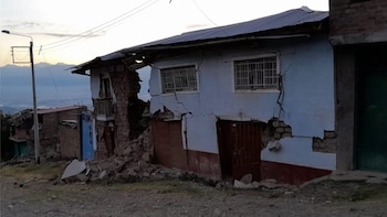 Vista exterior de una casa con paredes agrietadas y derrumbadas, escombros en el suelo, un poste de luz, y montañas a lo lejos bajo un cielo claro