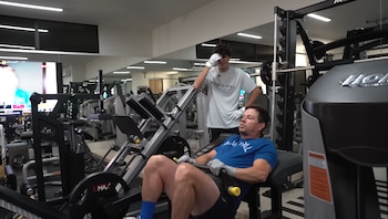 Mark Wahlberg está acostado en una máquina de prensa de piernas en un gimnasio moderno, mientras un hombre más joven lo observa de pie