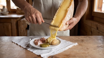 Una persona raspa queso fundido desde una rueda hacia un plato con patatas, pepinillos y embutido sobre una mesa de madera.