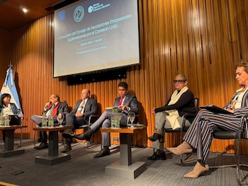 Seis personas sentadas en un panel sobre un escenario con una pantalla de proyección. Hay atriles individuales con vasos y botellas de agua y una bandera de Argentina
