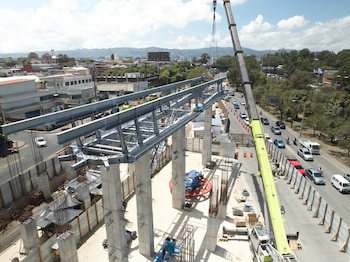 Grúa de gran tamaño eleva estructura metálica sobre pilares de concreto de una construcción. Debajo, calle con vehículos, conos de tráfico, edificios y montañas