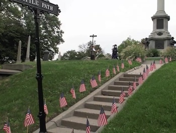 Escaleras de hormigón flanqueadas por pequeñas banderas de EE. UU. en un cementerio con césped verde, árboles, y un gran monumento de piedra al fondo. Un cartel negro dice "BATTLE PATH"