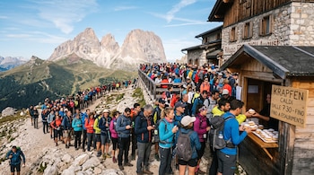 Una larga fila de personas ante un puesto de madera con un cartel de krapfen junto a un refugio de piedra con montañas de fondo.