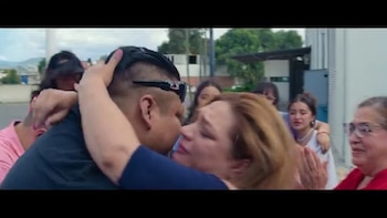 Primer plano de un hombre y una mujer abrazándose. Detrás de ellos, otras personas. Al fondo, edificios y el cielo visible